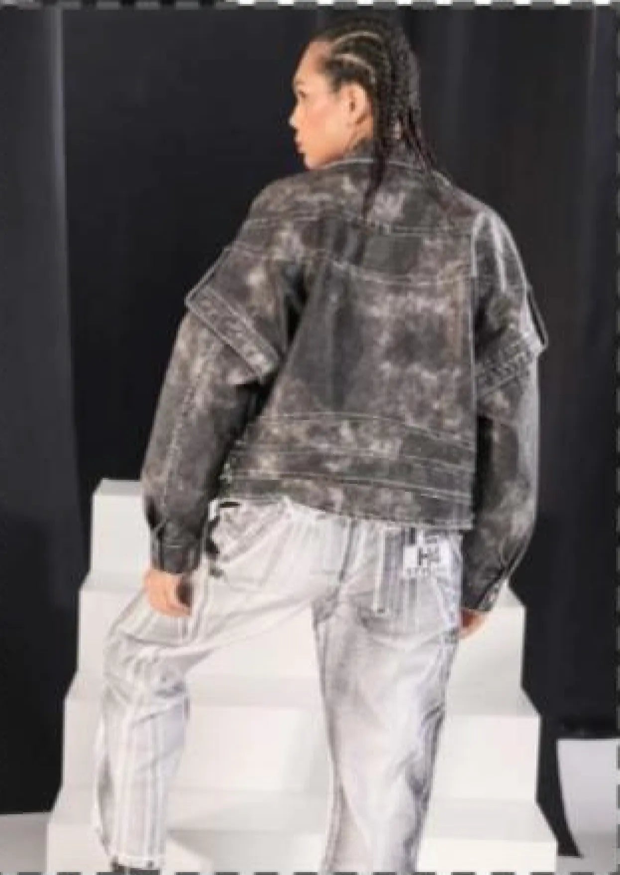 Person wearing a distressed black jacket and light gray pants on a plain background